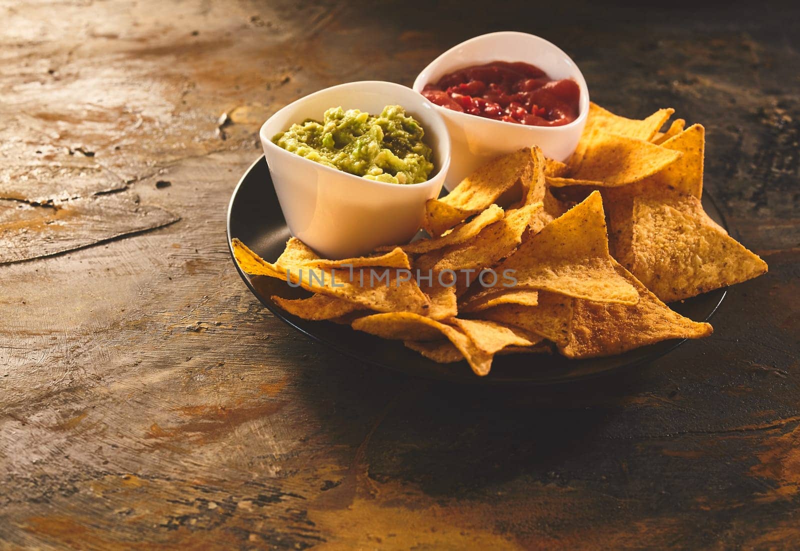 Single serving of delicious crunchy appetizer of triangular yellow corn tortilla chips beside little bowls of guacamole and salsa dip