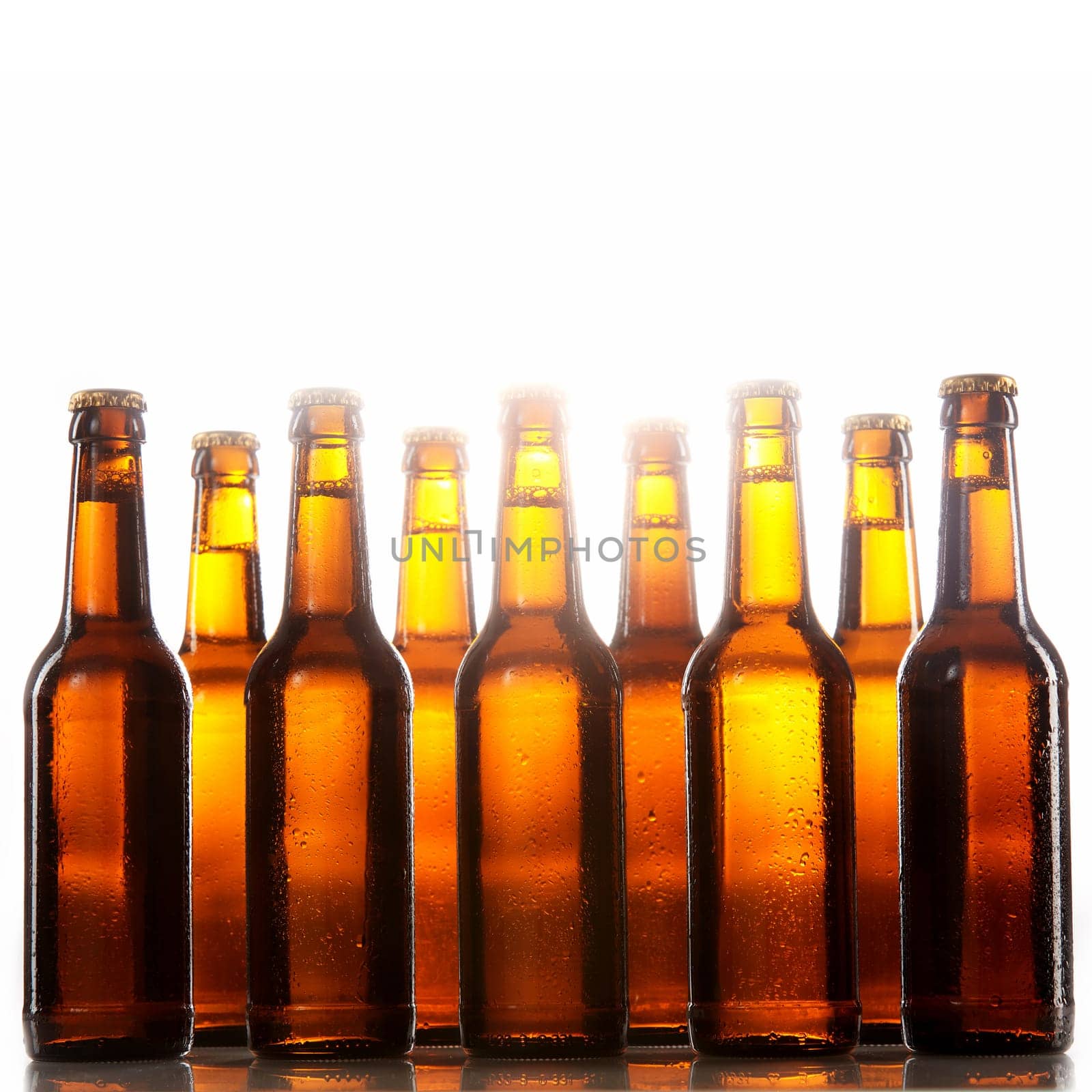 Tall beer bottles with no labels and metal caps stand two rows deep against a white background
