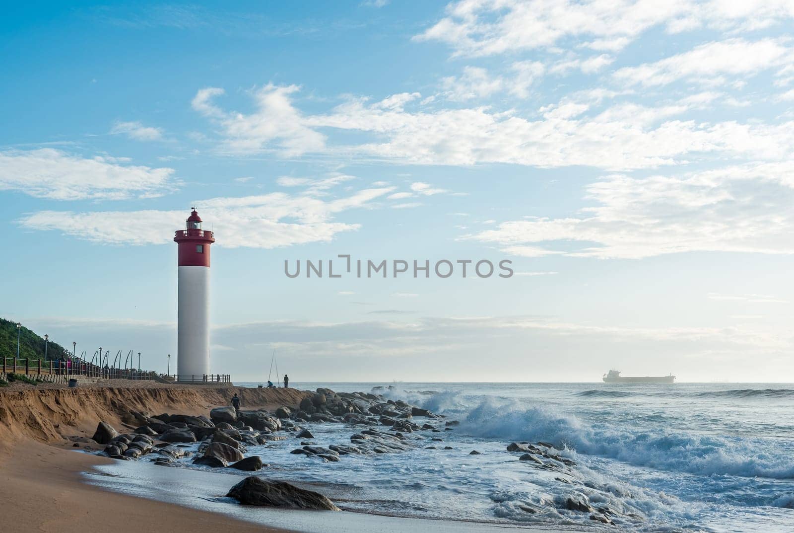 DURBAN, SOUTH AFRICA - MARCH 12, 2016: Umhlanga Rocks Lighthouse with fishermen and people walking on the promenade. Ship on the Indian Ocean in the background
