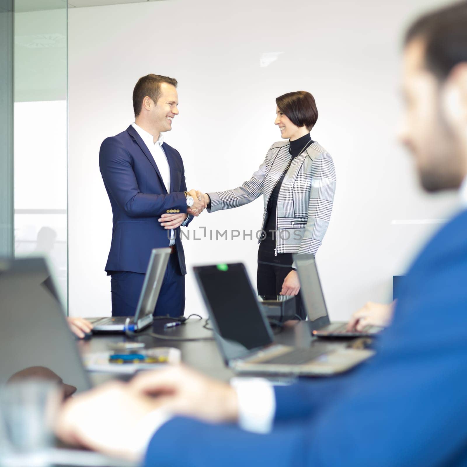Sealing a deal. Business people shaking hands, finishing up meeting in corporate office. Businessman working on laptop in foreground. Business and entrepreneurship concept.