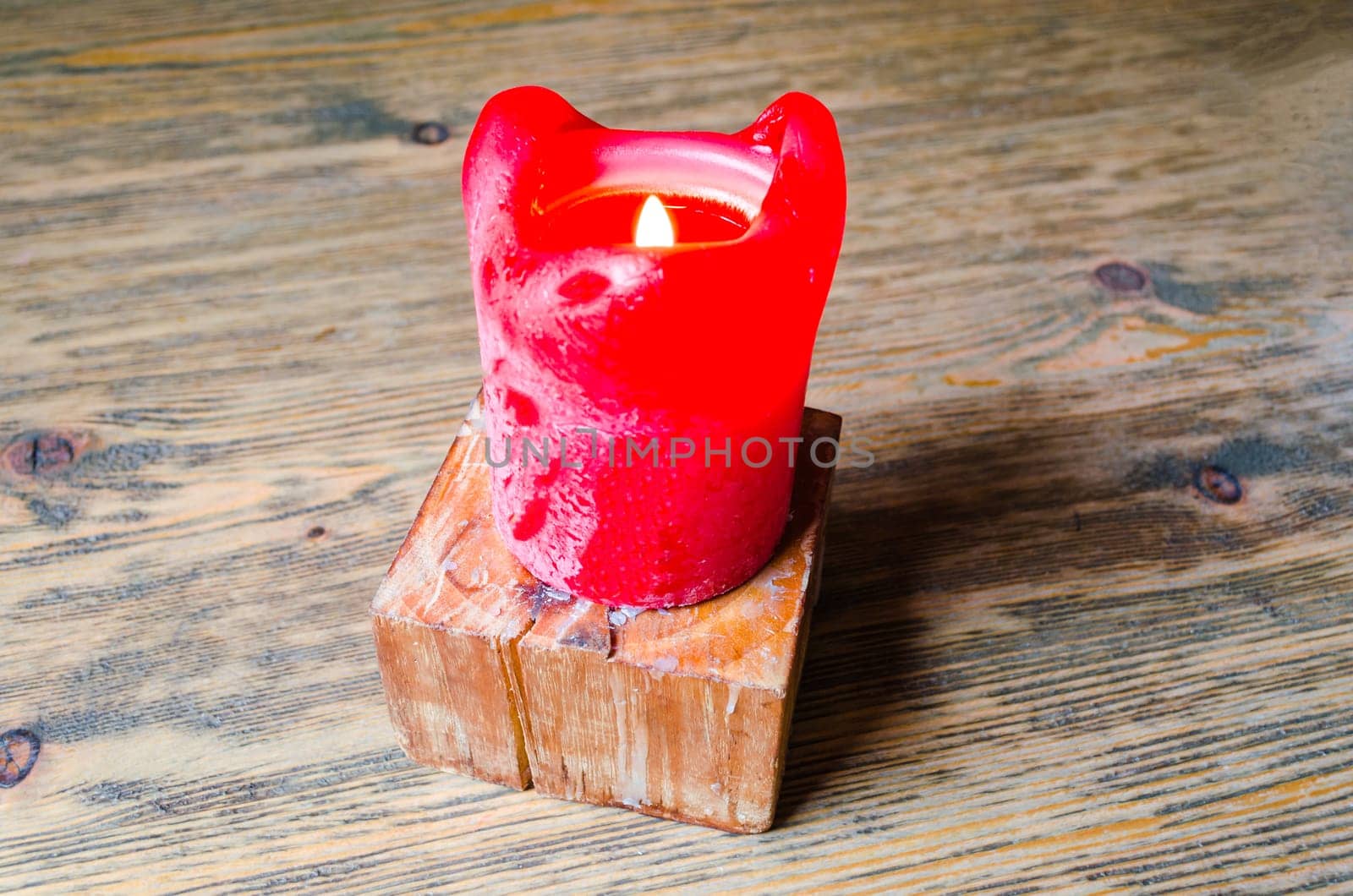 A single burning red candle on a table. Stands on a wooden candle stand.