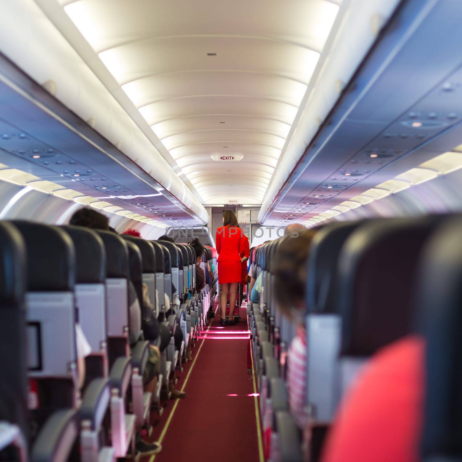 Interior of airplane with passengers on seats and stewardess in red uniform walking the aisle.