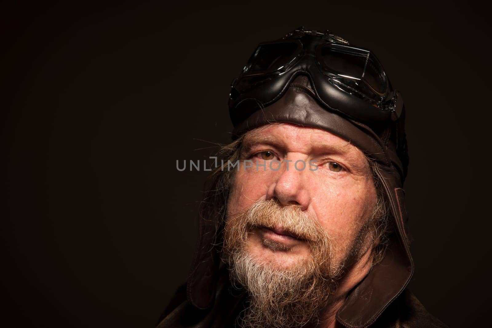 lateral head-and-shoulder portrait of an older man with a full bee,leather jacket and motorcycle goggles looking seriously into the camera