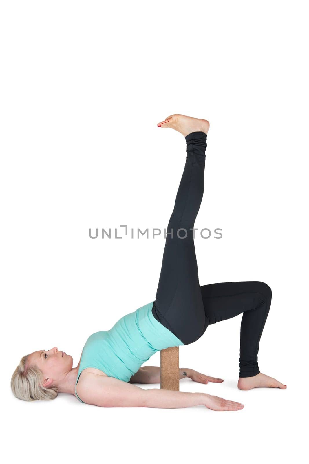 Full body view of a young blonde woman during yoga exercise "half bridge with her leg stretched upwards"" against a white background with a slight shadow. Wooden block as assistance under the buttocks."