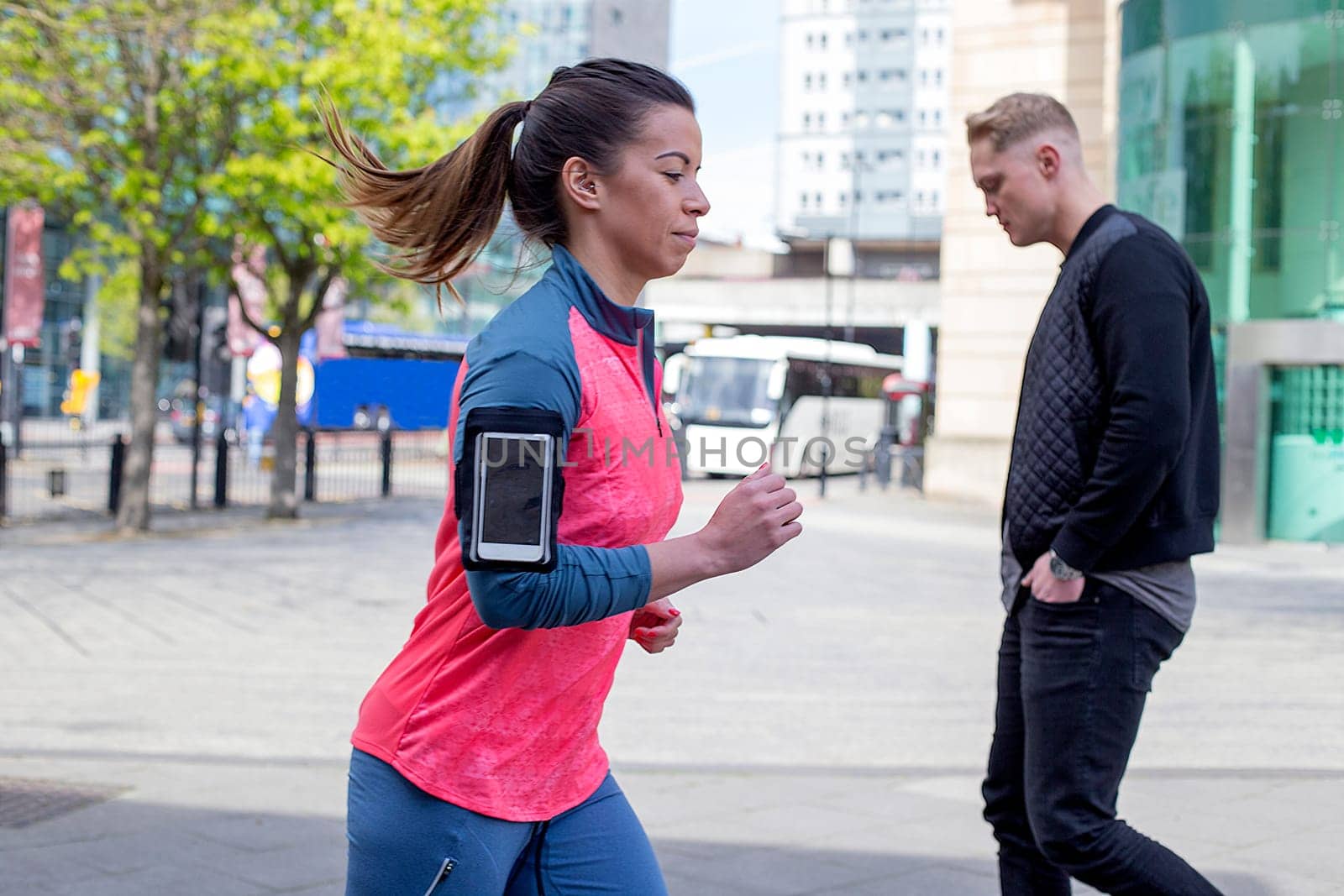 Woman running in the city. She has a smartphone in her arm strap. There is a man walking behind her.