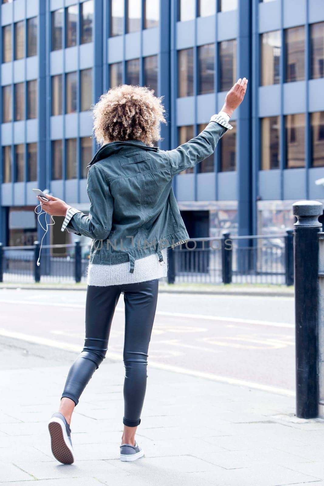 Young woman signalling for a taxi in the city centre. She is holding a smartphone with headphones and has her hand in the air.