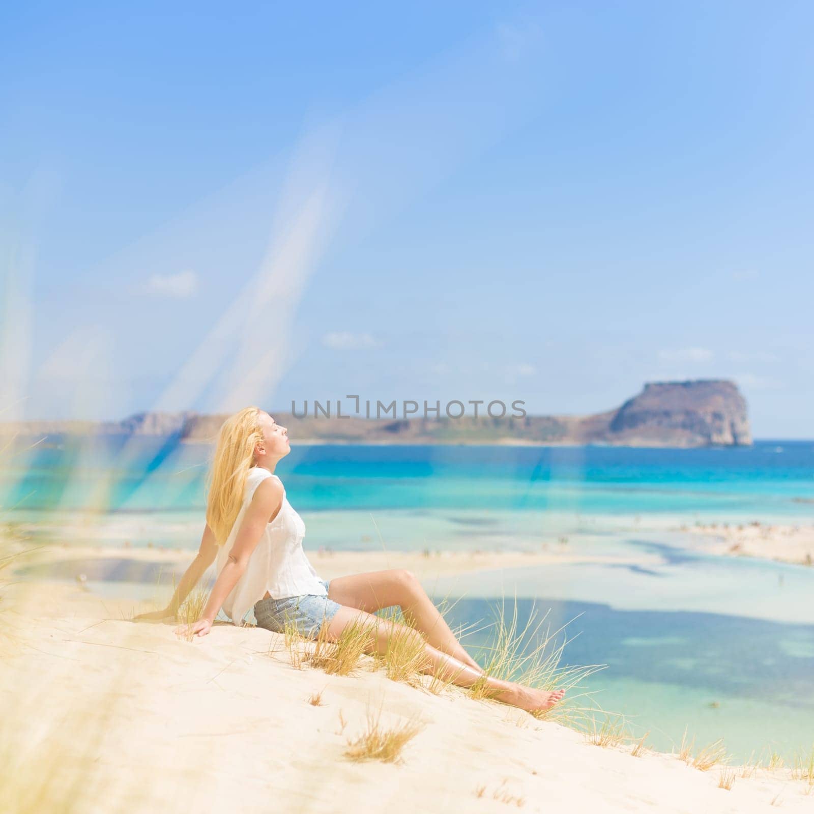 Relaxed woman enjoying sun, freedom and life an a beautiful sandy beach of Balos in Greece. Young lady feeling free, relaxed and happy. Vacations, freedom, happiness, enjoyment and well being.