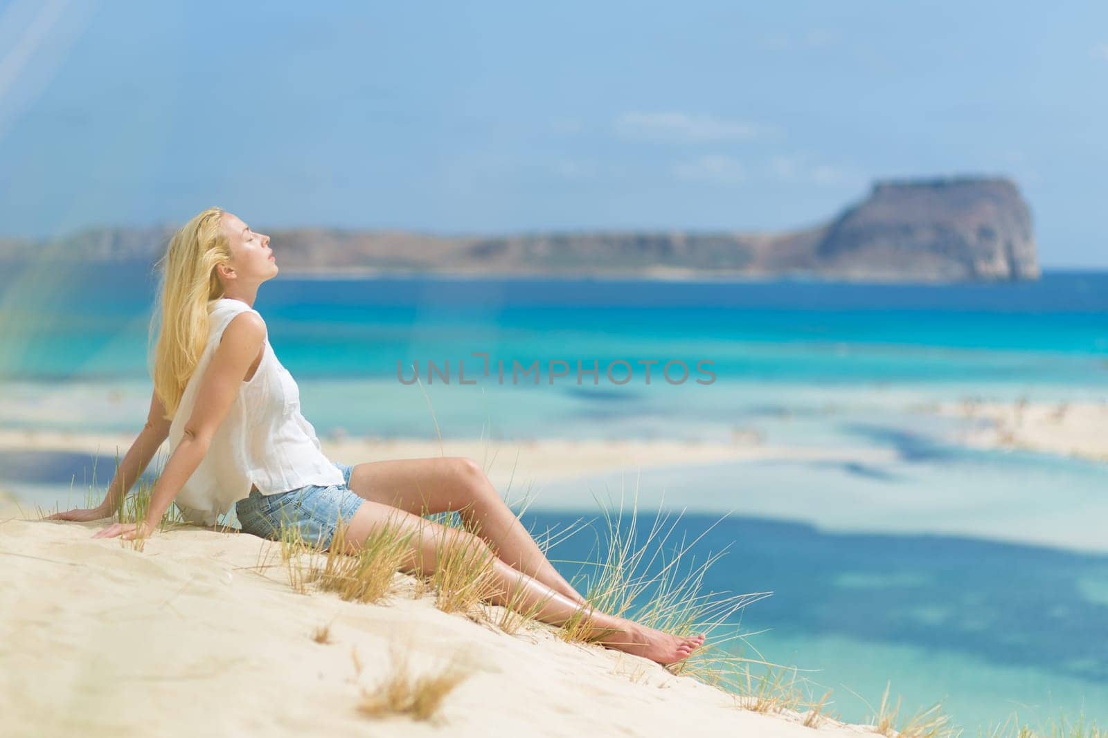Relaxed woman enjoying sun, freedom and life an a beautiful sandy beach of Balos in Greece. Young lady feeling free, relaxed and happy. Vacations, freedom, happiness, enjoyment and well being.