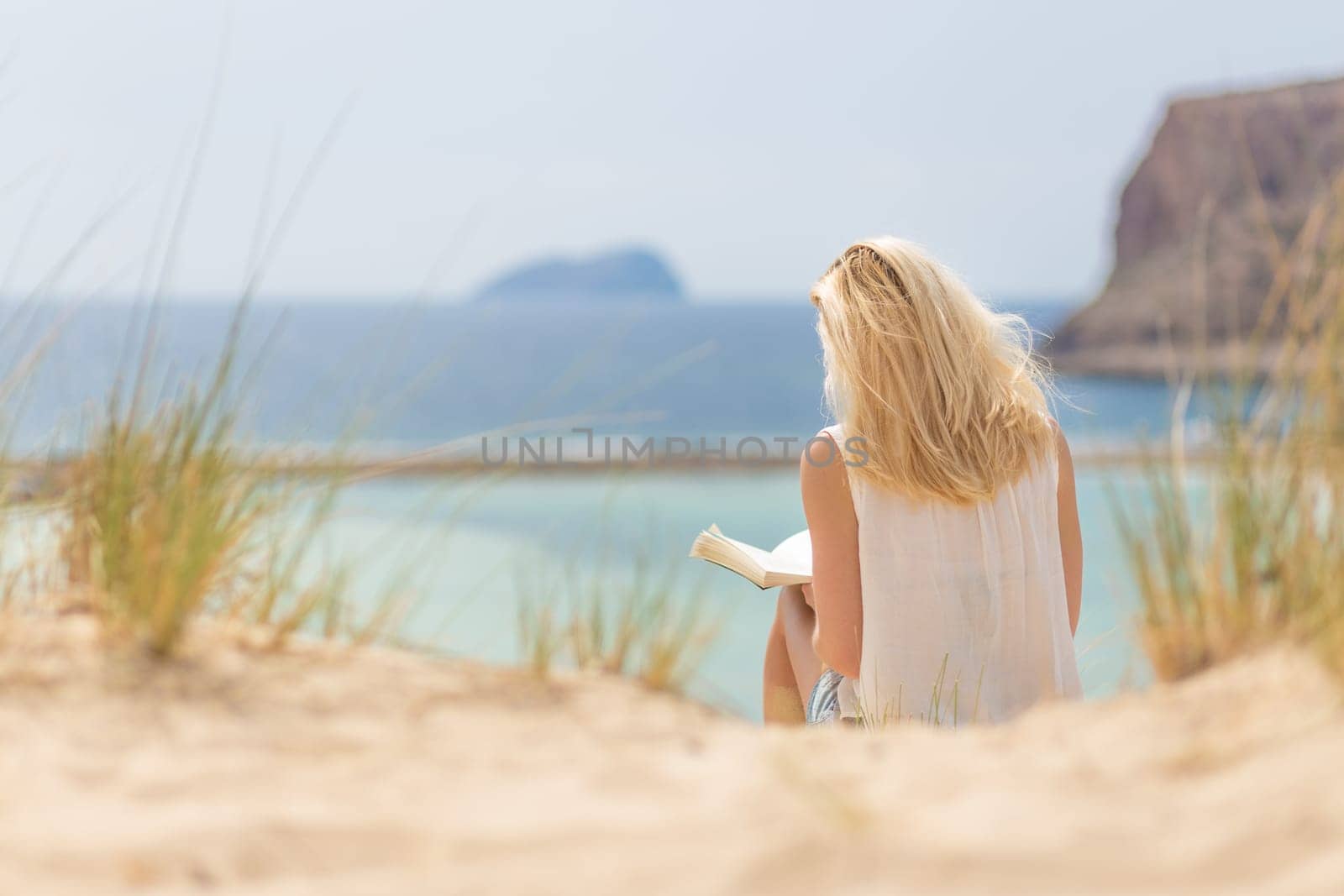 Relaxed woman enjoying sun, freedom and good book an beautiful sandy beach of Balos in Greece. Young lady reading, feeling free and relaxed. Vacations, freedom, happiness, enjoyment and well being.