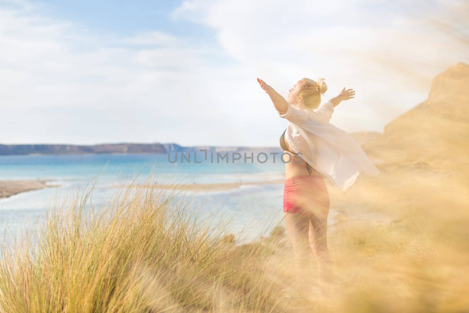 Relaxed woman in white shirt, arms rised, enjoying sun, freedom and life an beautiful beach. Young lady feeling free, relaxed and happy. Concept of vacations, freedom, happiness, joy and well being.