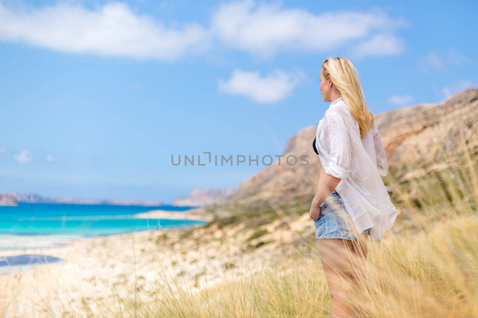Relaxed woman in white shirt looking at distance, enjoying beautiful nature, freedom and life at serene landscape at Balos beach, Greece. Concept of vacations, freedom, happiness, joy and well being.