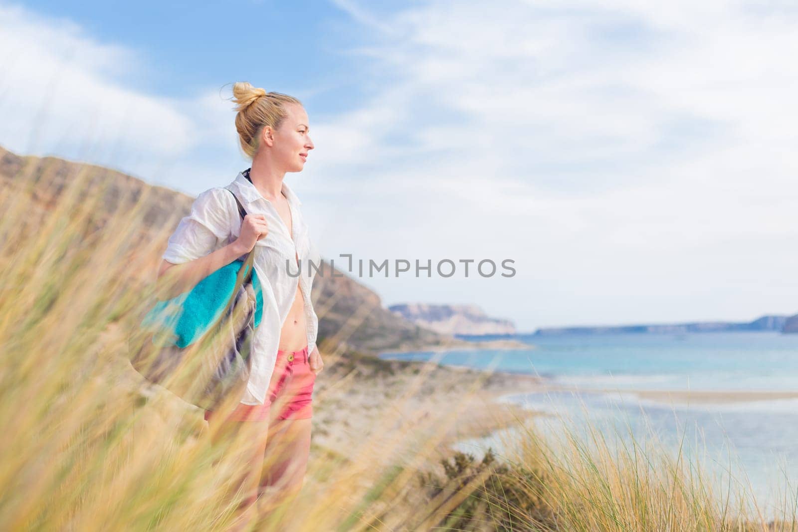 Relaxed woman on vacations in white loose shirt carrying beach bag and towel, enjoying beautiful coastline view of white sandy lagoon at Balos beach, Greece.