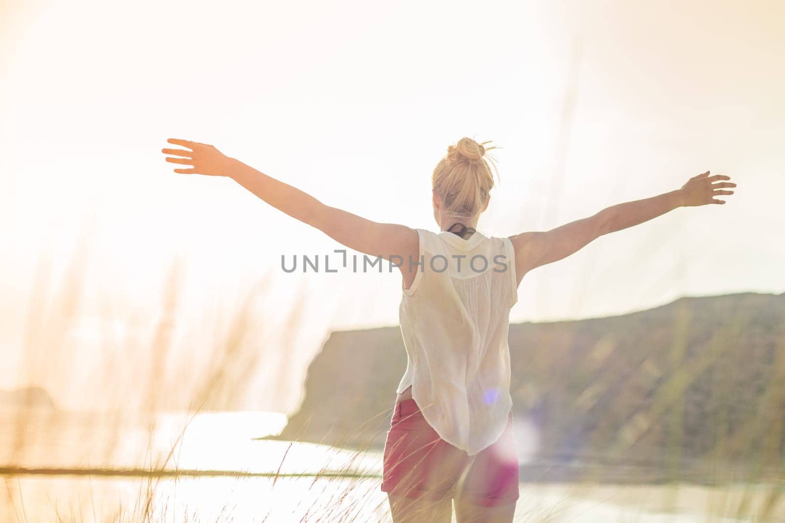 Relaxed woman in white shirt, arms rised, enjoying sun, freedom and life an beautiful beach. Young lady feeling free, relaxed and happy. Concept of vacations, freedom, happiness, joy and well being.
