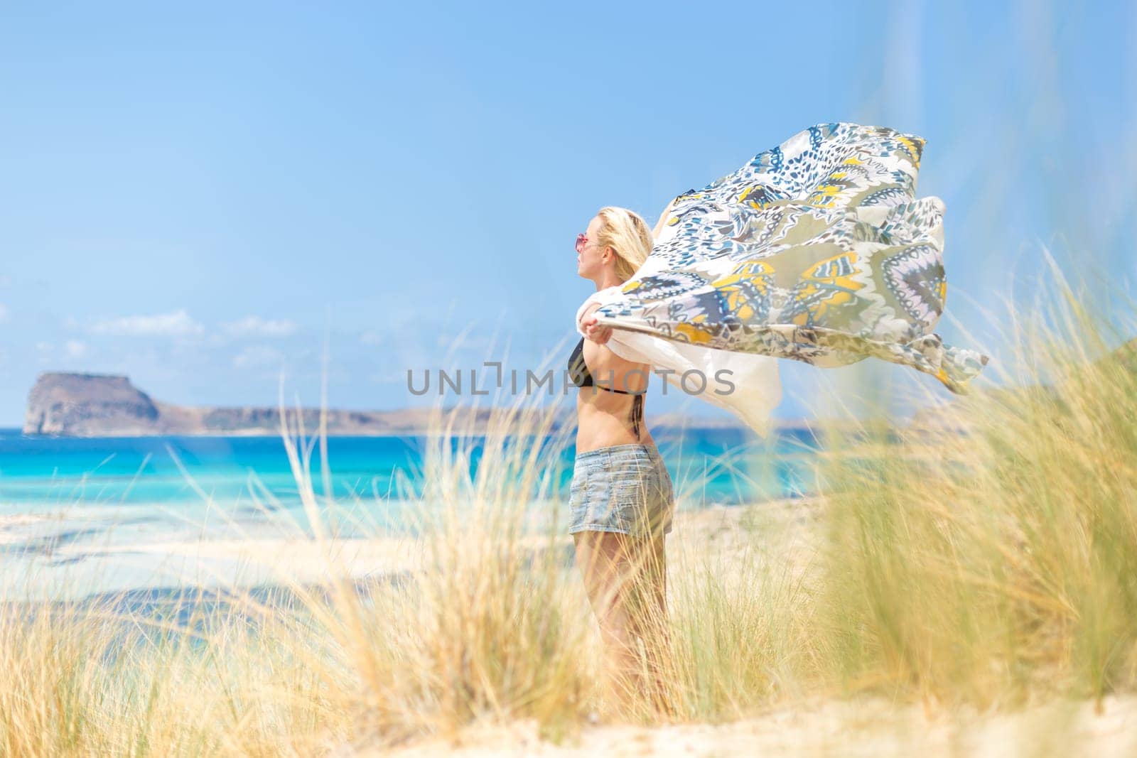Relaxed woman, arms rised, holding colorful scarf, enjoying sun, freedom and life at beautiful beach. Young lady feeling free, relaxed and happy. Concept of vacations, freedom, joy and well being.