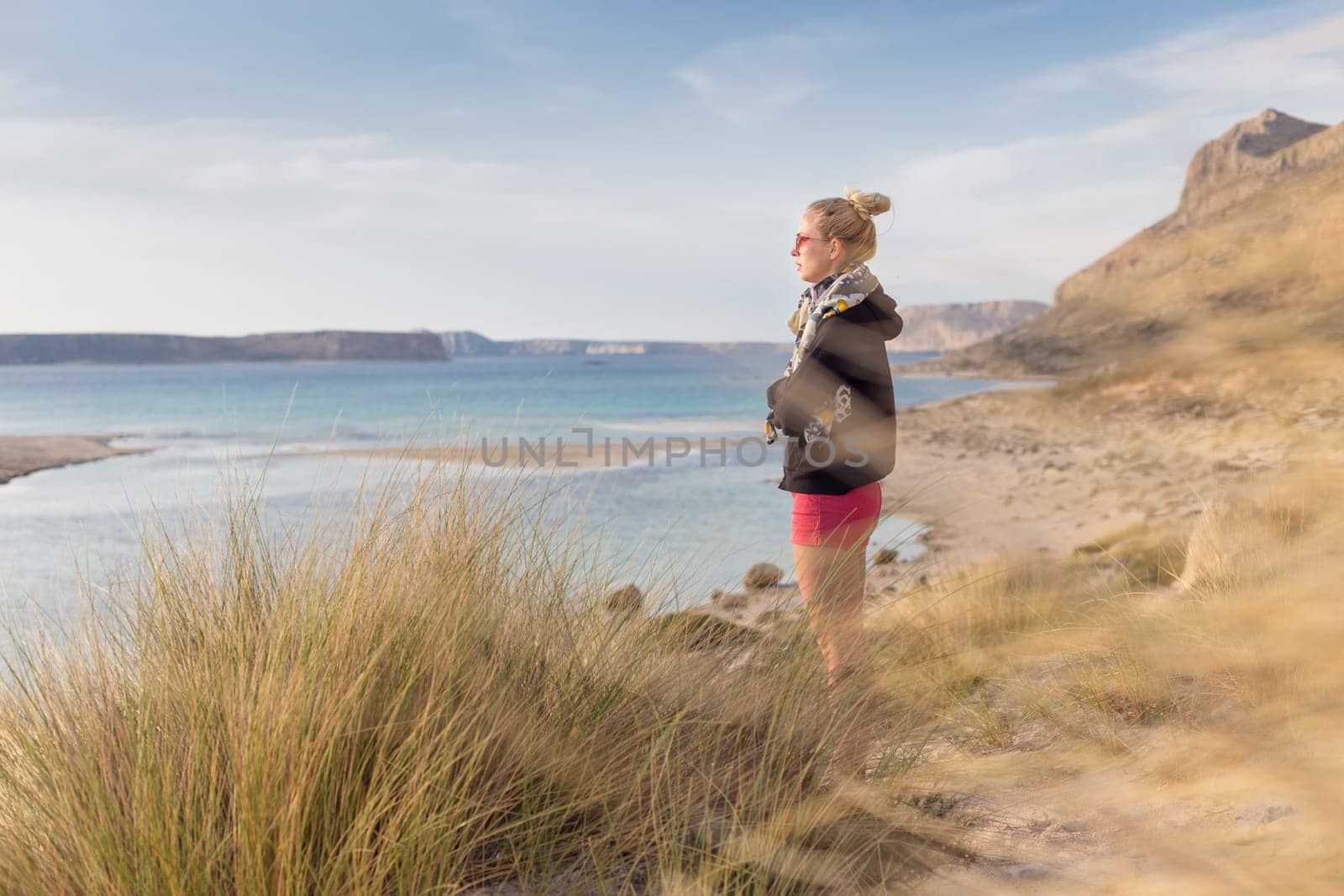 Relaxed woman in dark hoodie and colorful scarf looking at distance, enjoying beautiful nature, freedom and life at serene landscape at Balos beach, Greece. Concept of well being.
