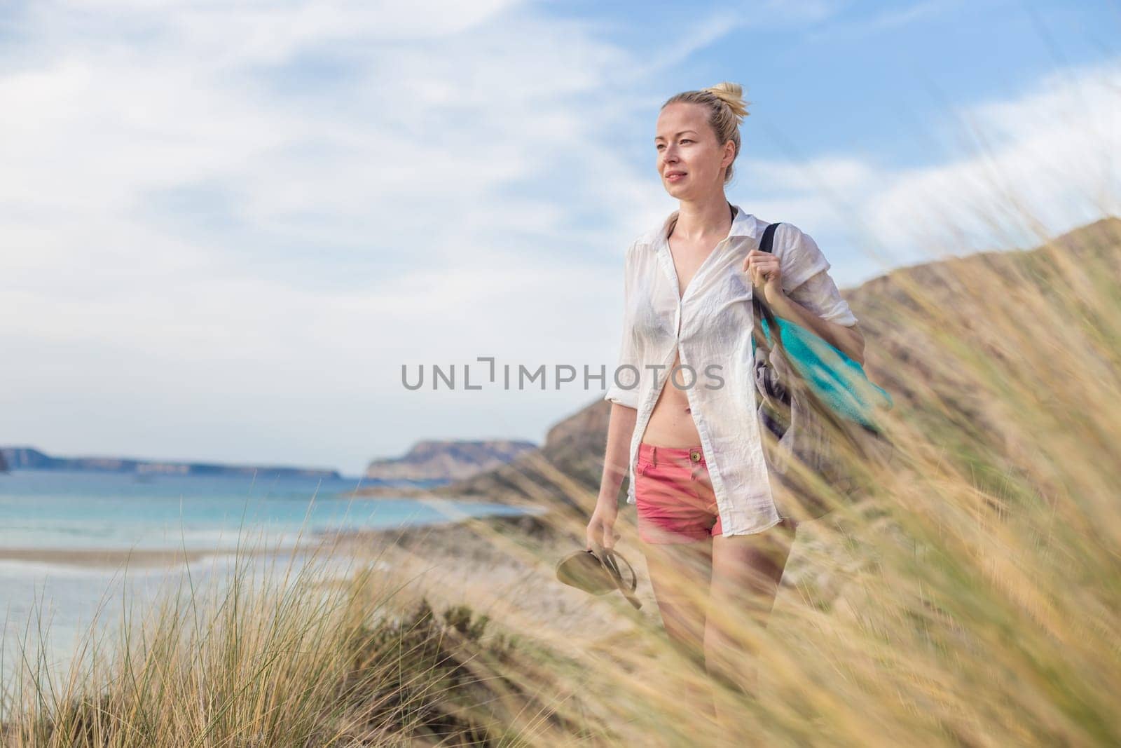 Relaxed woman on vacations in white loose shirt carrying beach bag and towel, enjoying beautiful coastline view of white sandy lagoon at Balos beach, Greece.