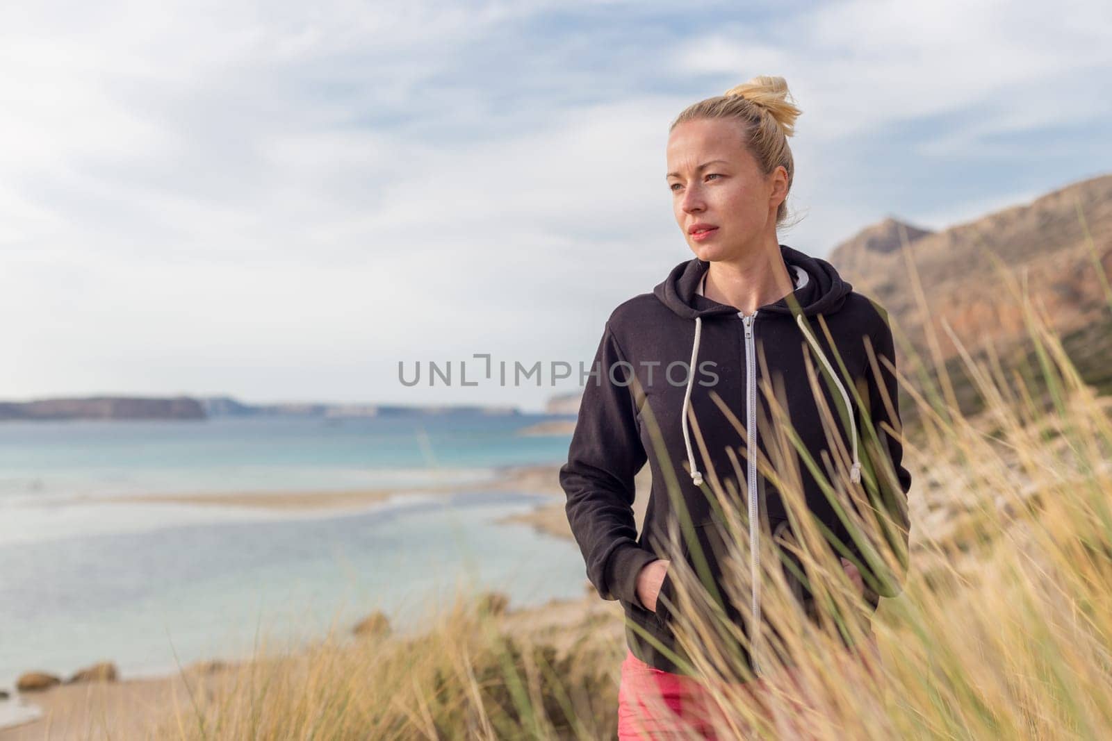 Relaxed woman in dark hoodie and colorful scarf looking at distance, enjoying beautiful nature, freedom and life at serene landscape at Balos beach, Greece. Concept of well being.