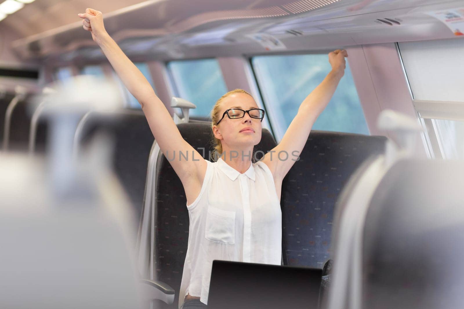 Businesswoman traveling by train, streching her upper body arms reised taking a break from laptop work. Business travel concept.