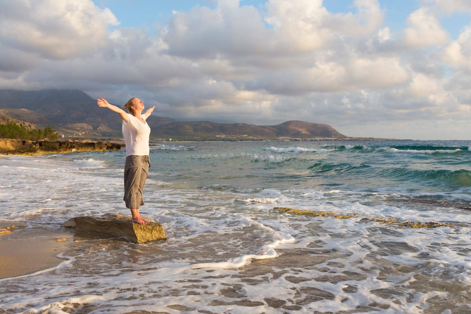 Relaxed woman, arms rised, enjoying sun, freedom and life an beautiful beach in sunset. Young lady feeling free, relaxed and happy. Concept of vacations, freedom, happiness, enjoyment and well being.