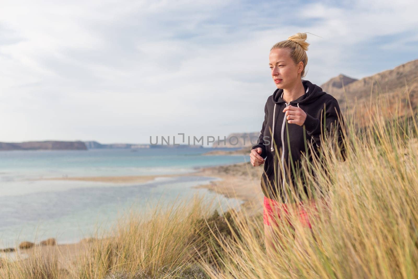 Active woman in dark hoodie running on serene Balos beach, Greece, enjoying beautiful nature and nice views. Healthy active lifestyle.