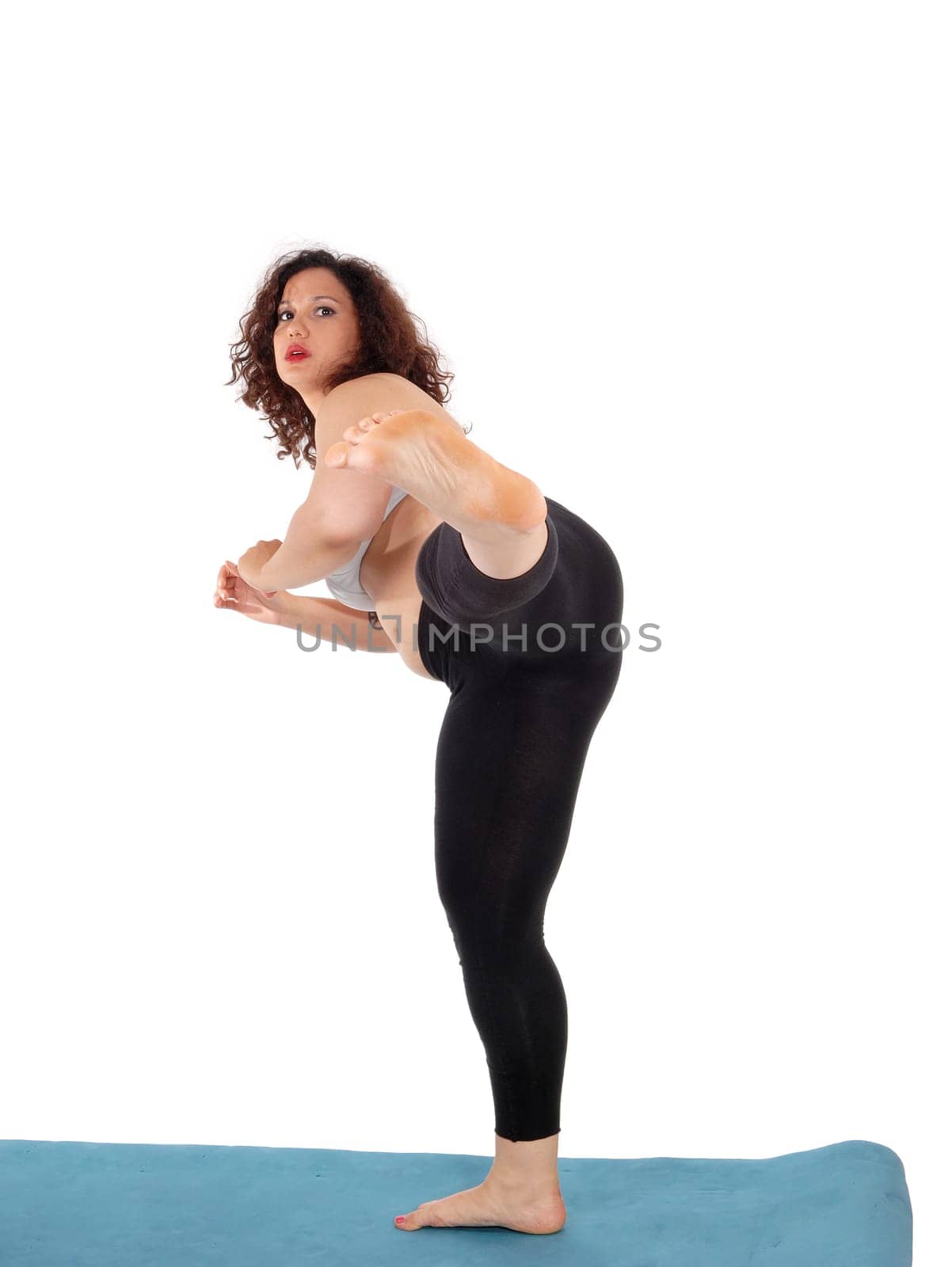 An exercising young woman in black pants standing on a blue mat,.kicking with her leg, isolated for white background..