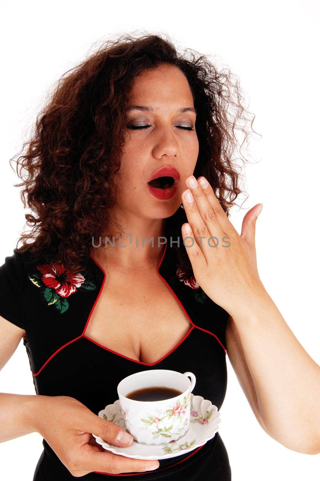 A really tired woman in a black dress holding the hand over her mouth,.yawning, with a cup of coffee, isolated for white background..