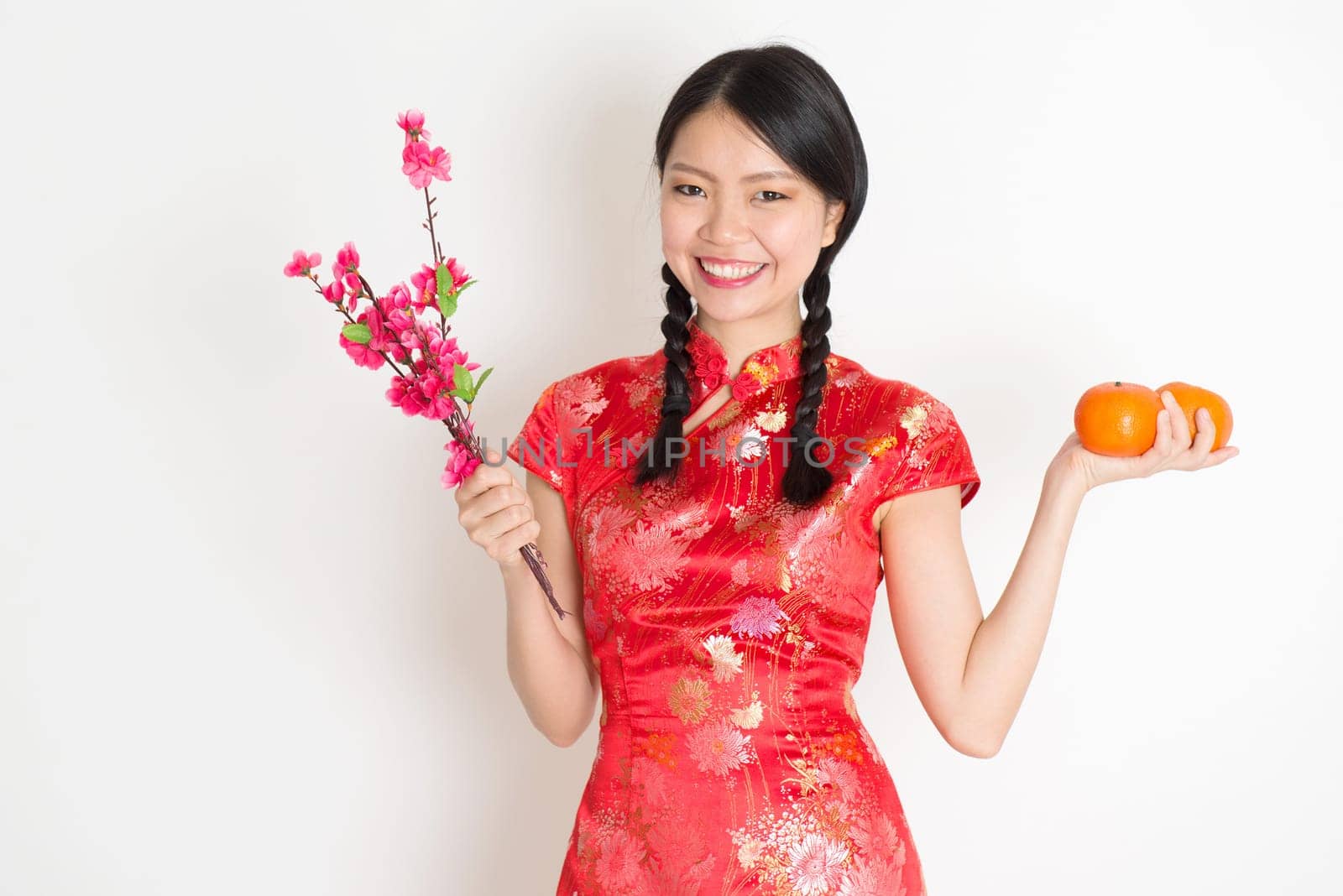Portrait of Asian Chinese girl hands holding tangerine orange and plum blossom, in traditional red cheongsam standing on plain background.