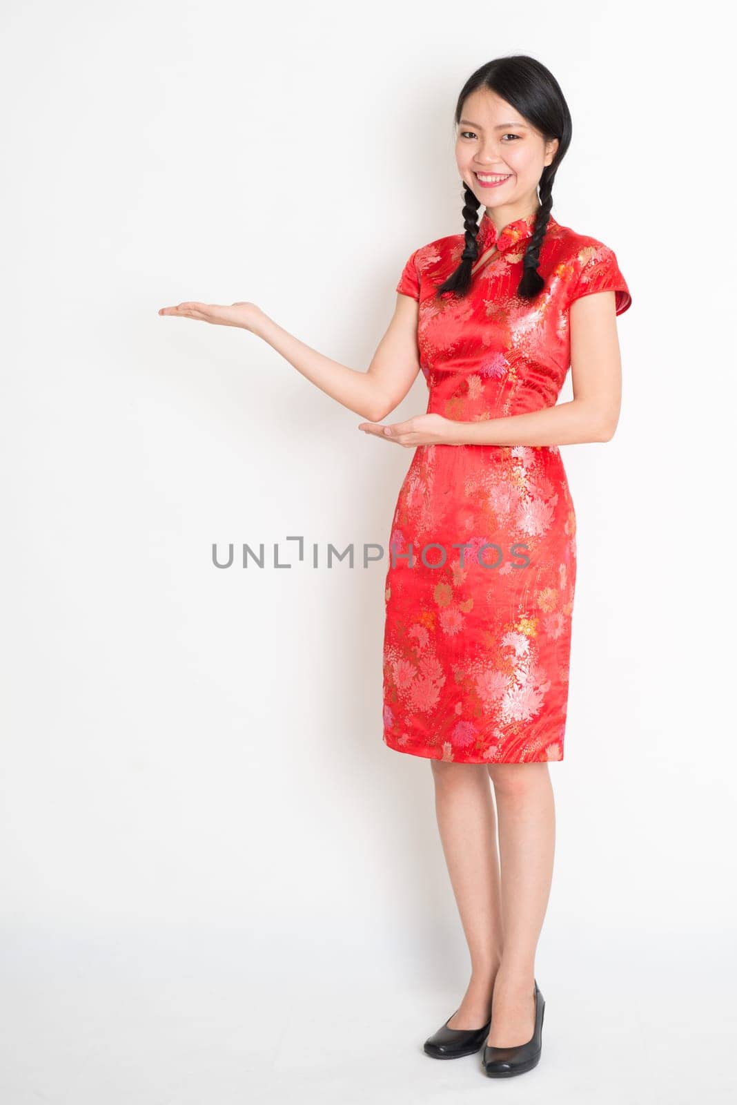 Portrait of full length Asian Chinese girl, hands showing something, in traditional red cheongsam standing on plain background.