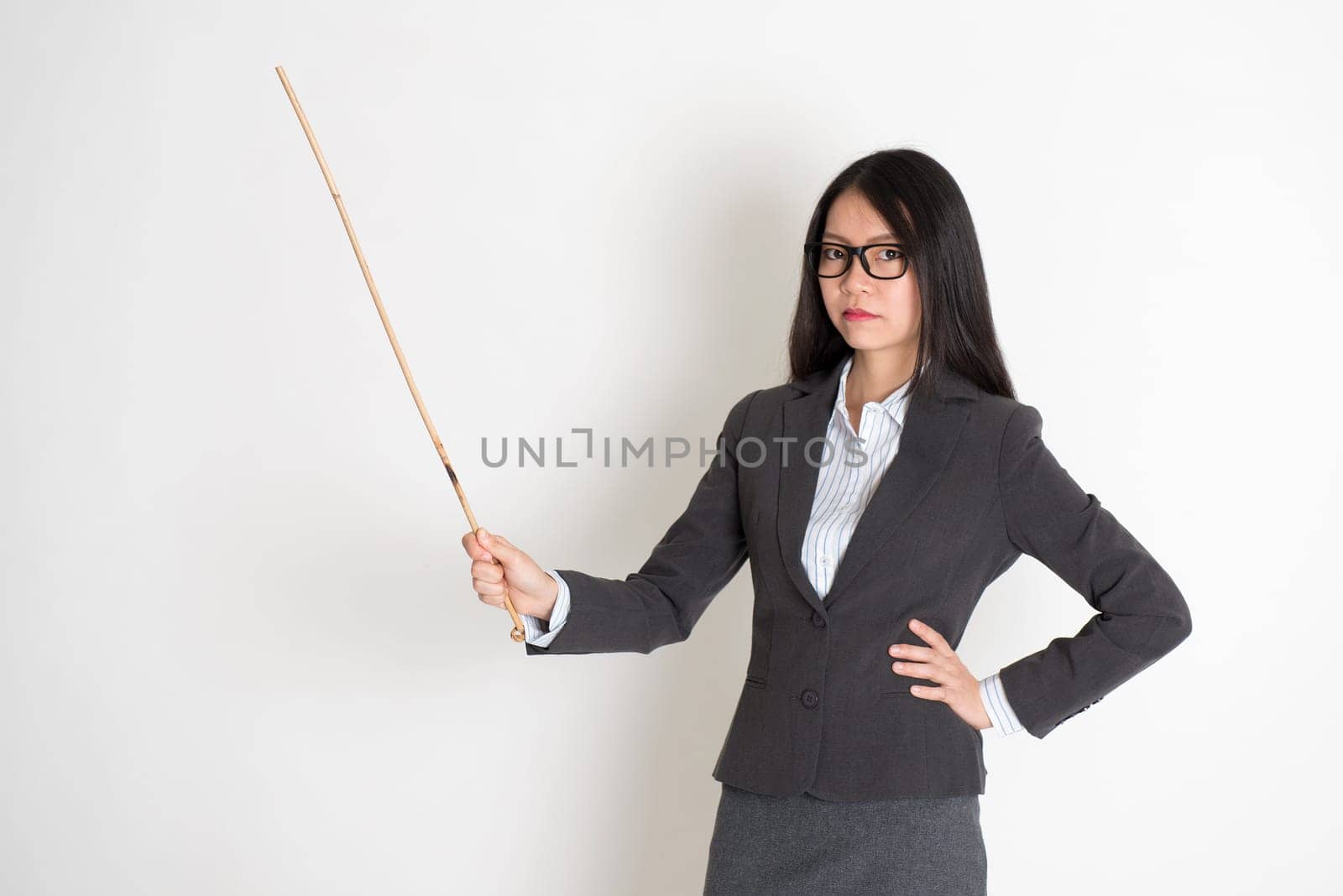Asian female teacher angry and holding a stick, standing on plain background.