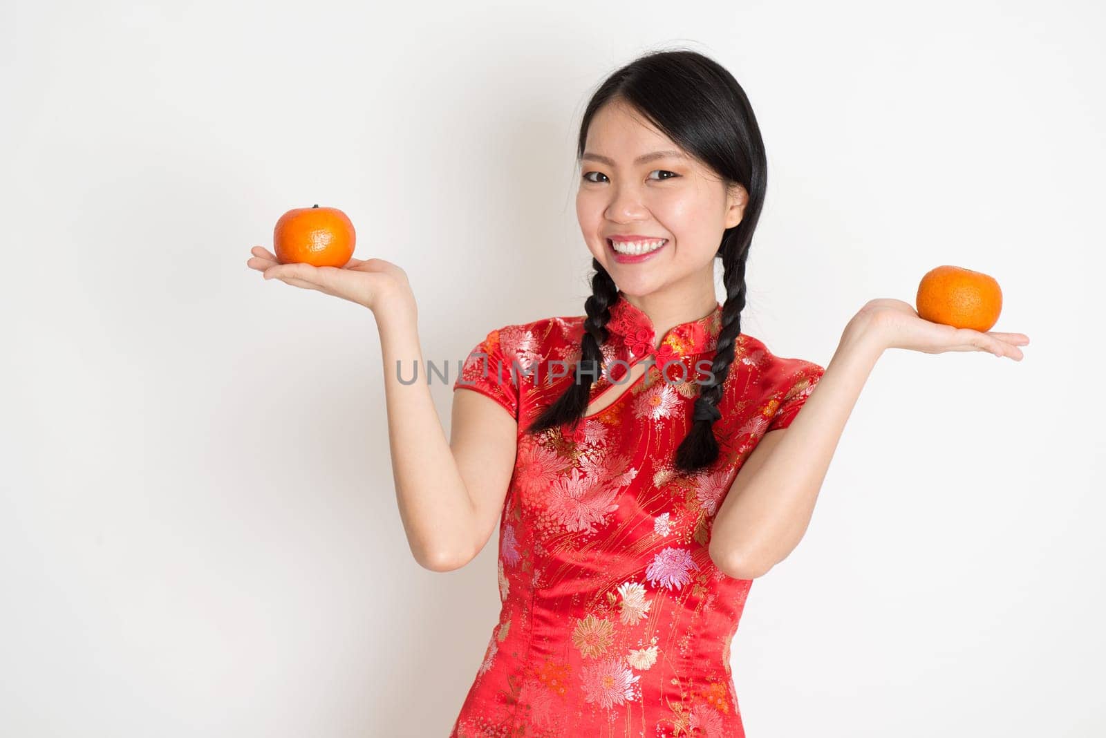 Portrait of Asian Chinese girl hands holding tangerine orange, in traditional red qipao standing on plain background.