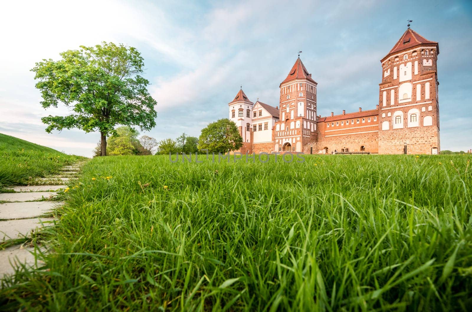 Green trees and grass and blue sky at sunny day. Stone road to beautiful Mir castle in Belarus. Medieval fort became sight for tourists.