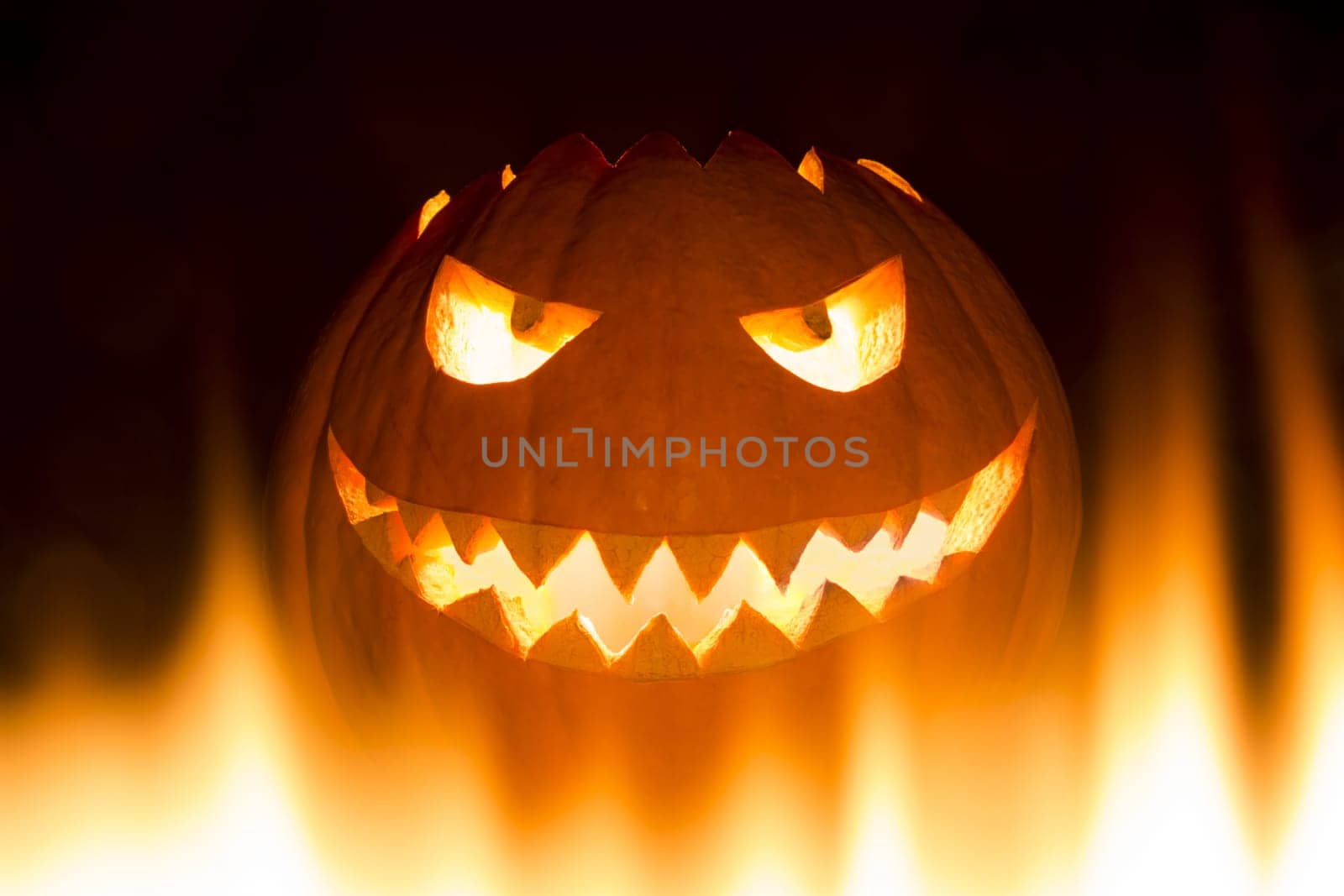 The big Helloween pumpkin has a face that laughs and is cheeky with shining eyes and a mouth with pointed teeth. Perspective from below in burning flames.