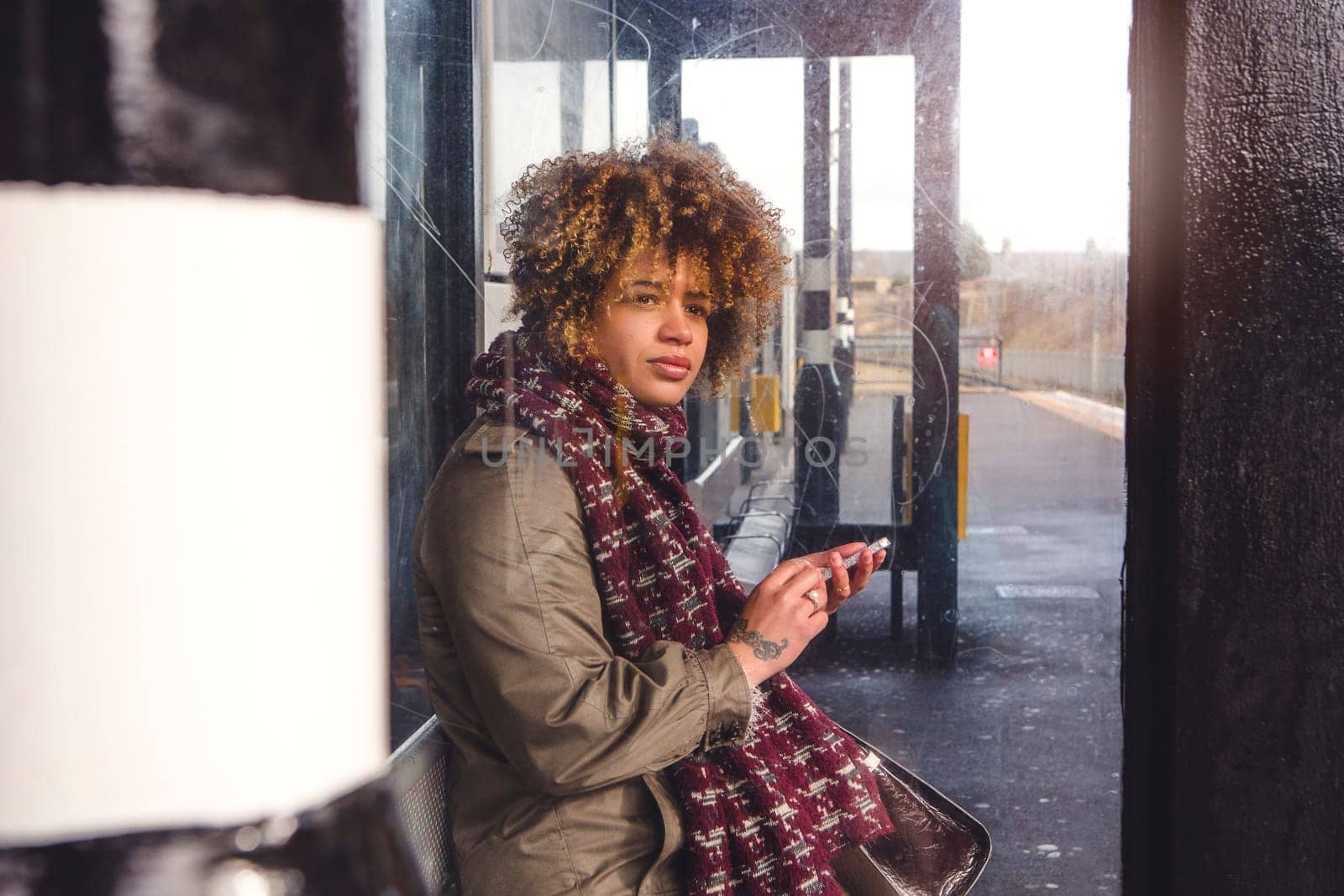 Woman using her smart phone as she waits for her train at the platform. She is sitting in the shelter, looking out for the train.