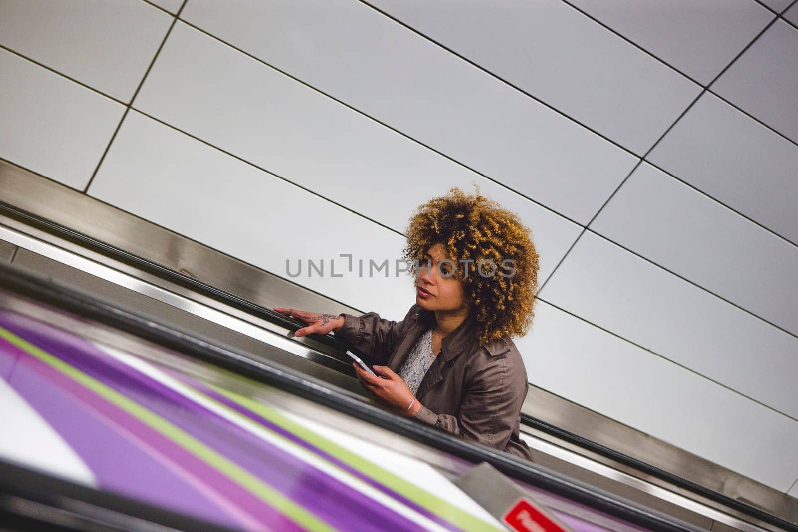 Woman travelling up an escalator from the subway train platform. She is holding a smart phone.