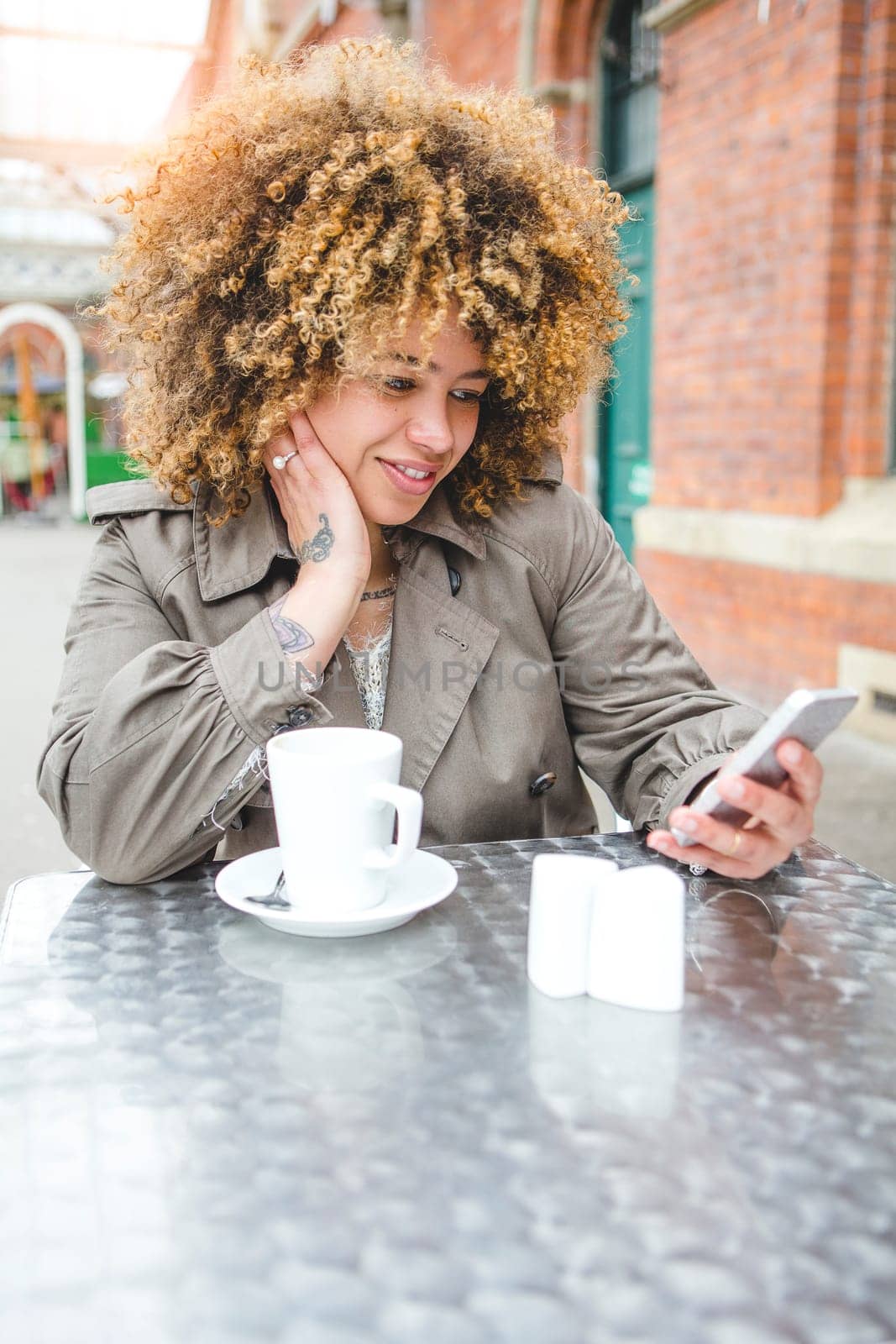 Woman sitting outdoors at a coffee shop with a drink. She is using a smart phone.