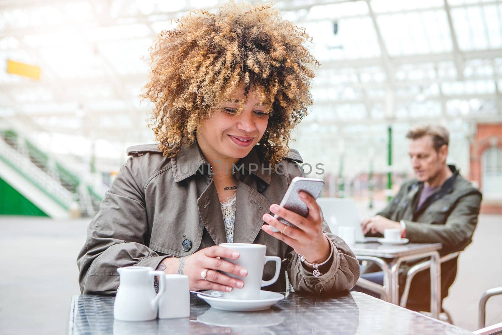 Cheerful woman using her smart phone at a cafe in the train station while she waits for her train. She has a cup of tea on the table.