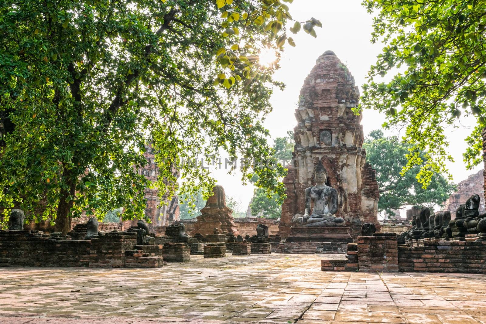 Buddha statue sitting position at front of pagoda under sun light surround by trees and ancient ruins of Wat Phra Mahathat temple in Phra Nakhon Si Ayutthaya Historical Park, Thailand