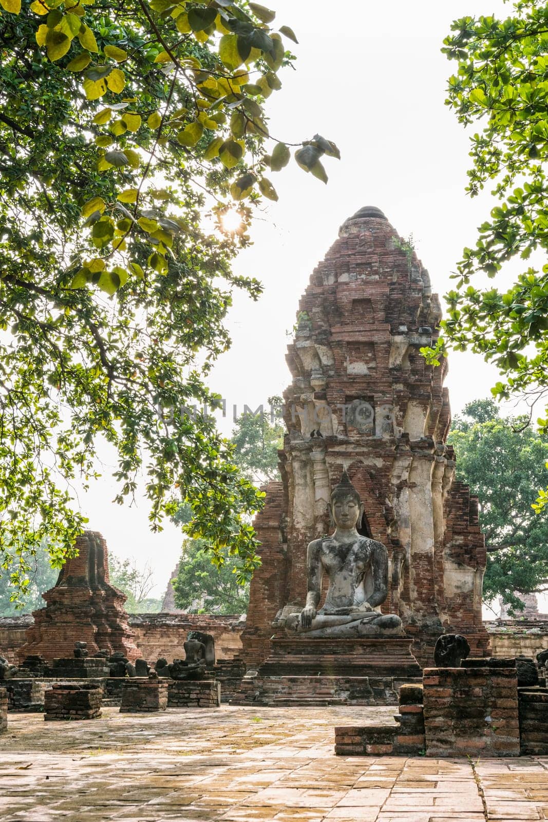 Buddha statue sitting position at front of pagoda under sun light surround by trees and ancient ruins of Wat Phra Mahathat temple in Phra Nakhon Si Ayutthaya Historical Park, Thailand