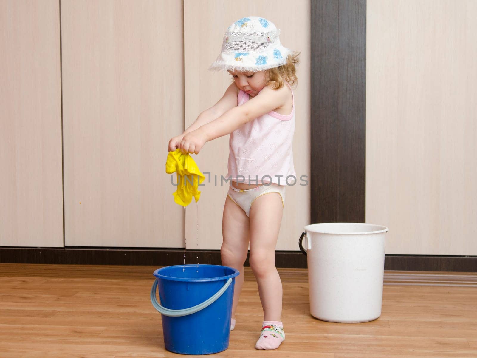 Two-year-old girl with pleasure and fun washing the floors in the room. Nearby is a small blue bucket. Girl washes the cloth in a pail.
