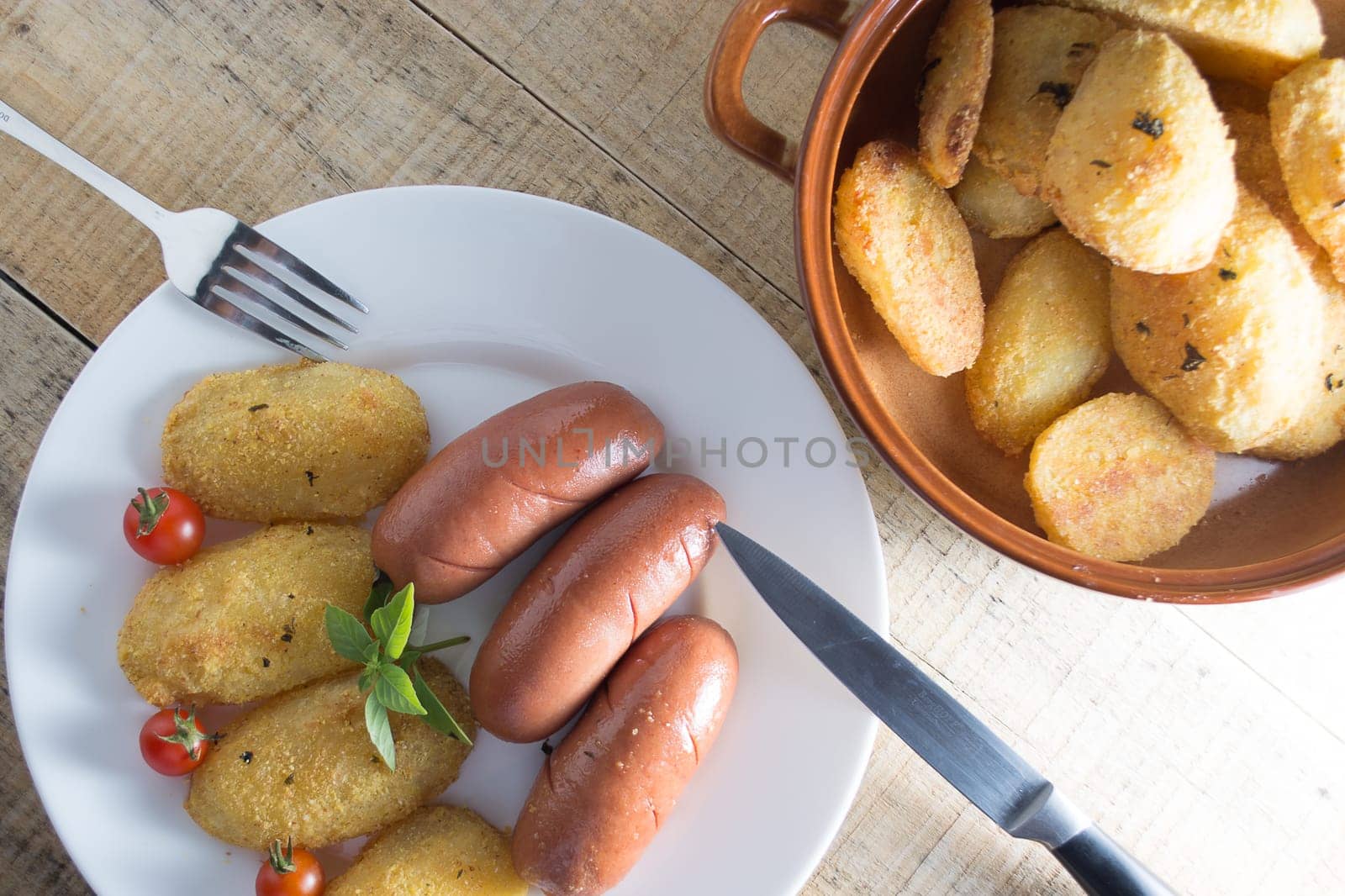 Pork sausages and breaded potatoes fried in a dish and decorated with cherry tomato.  Fork and knife placed next to dish on wooden table