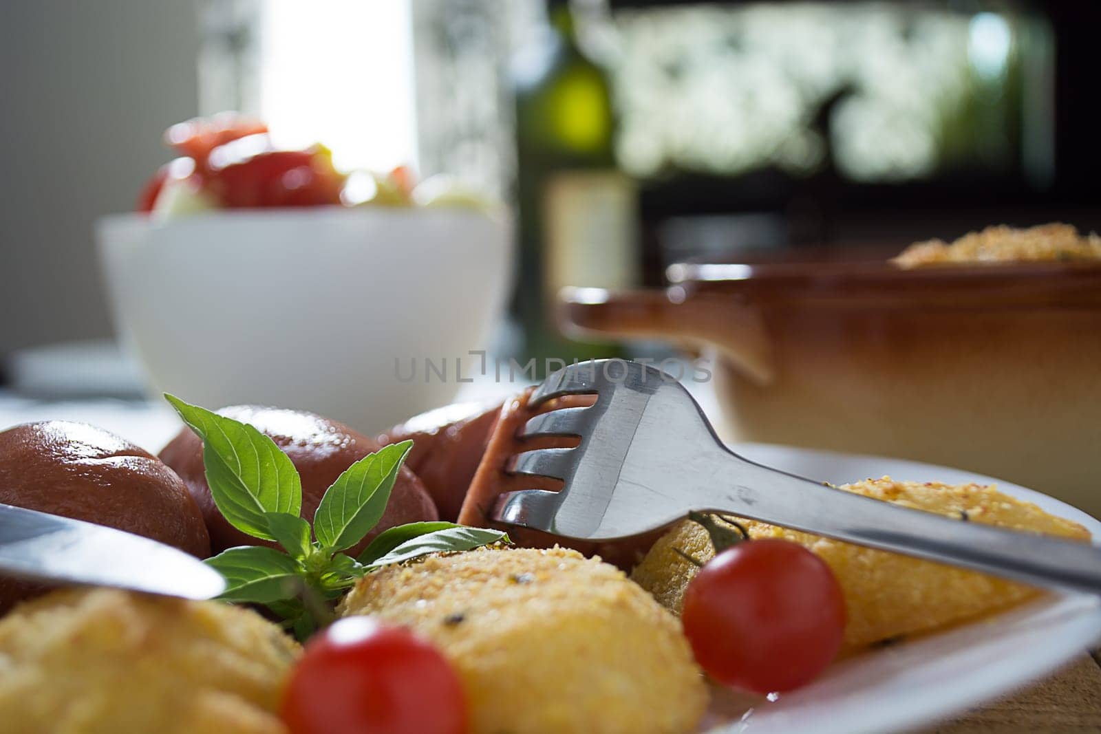 Pork sausages and breaded potatoes fried in a dish and decorated with cherry tomato. Fork and knife next to dish