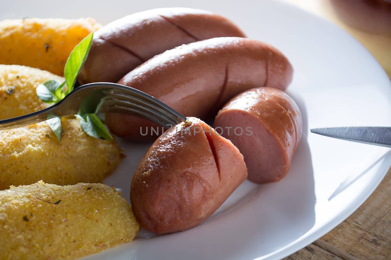 Pork sausages and breaded potatoes fried in a dish.  Fork and knife placed next to dish on wooden table