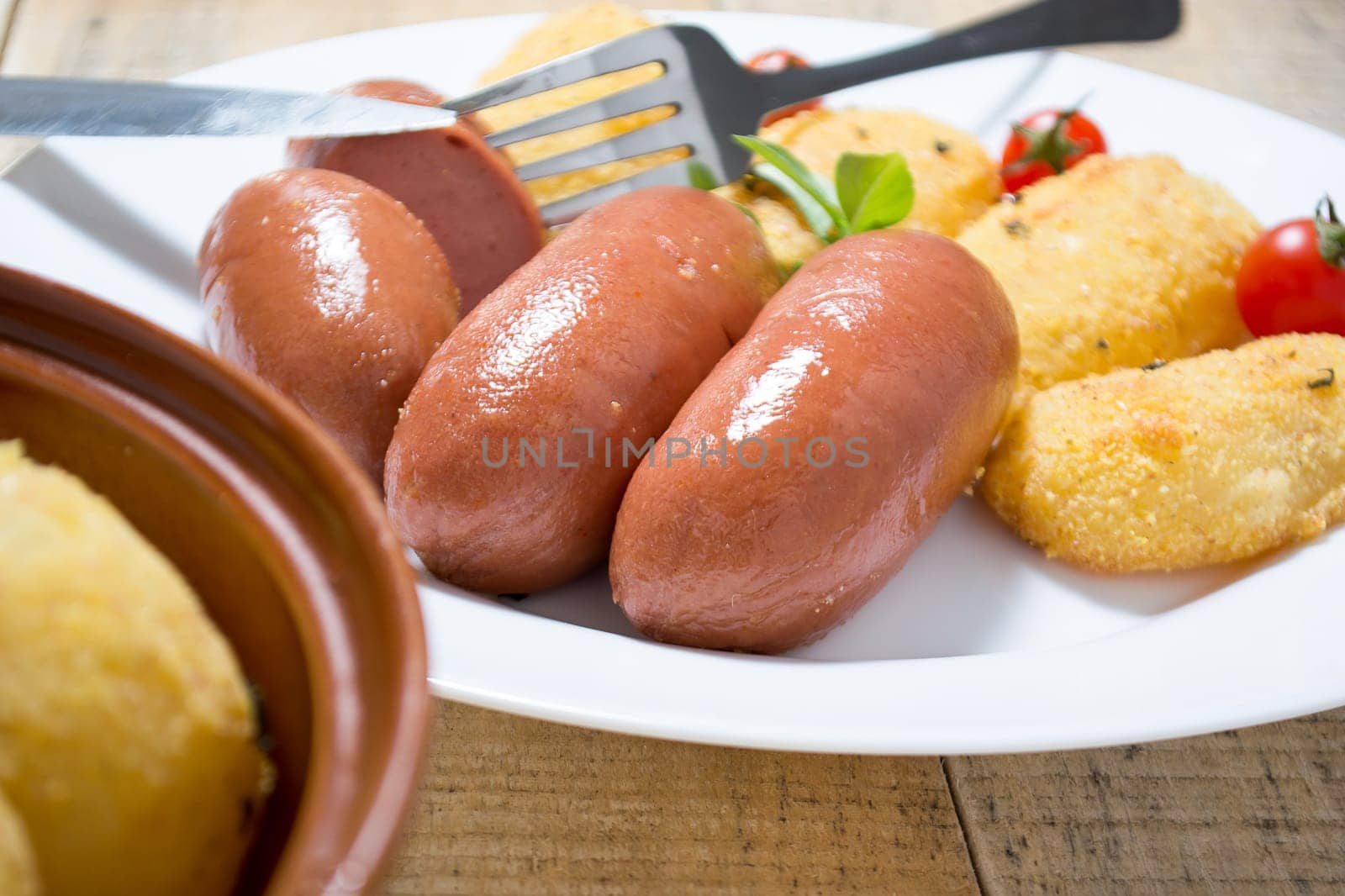 Pork sausages and breaded potatoes fried in a dish and decorated with cherry tomato.  Fork and knife placed next to dish on wooden table