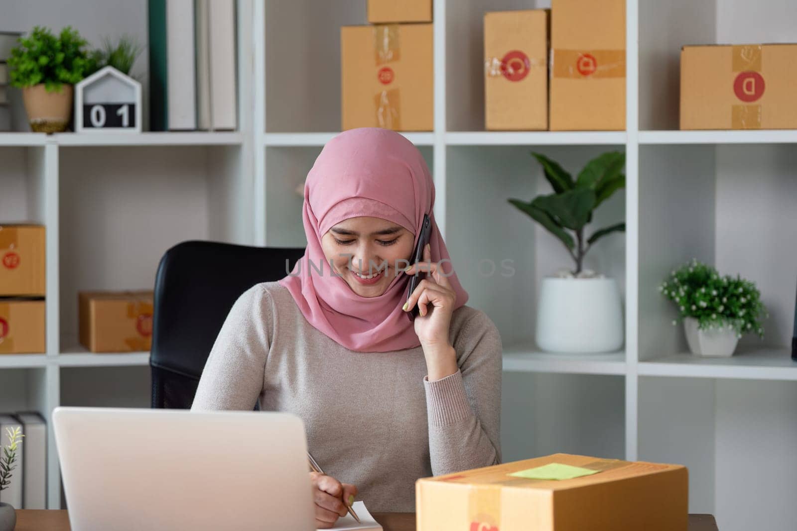 A smiling Muslim woman in a hijab speaks on the phone, overseeing her online business and packages in her home office.