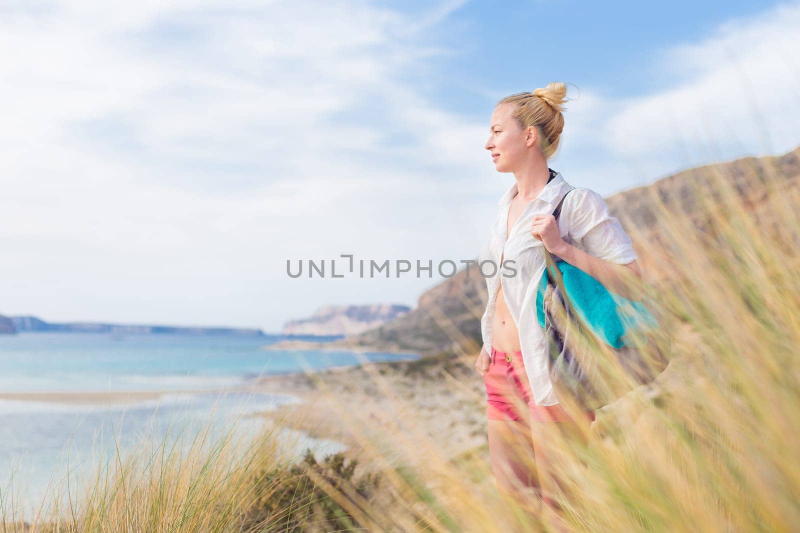 Relaxed woman on vacations in white loose shirt carrying beach bag and towel, enjoying beautiful coastline view of white sandy lagoon at Balos beach, Greece.