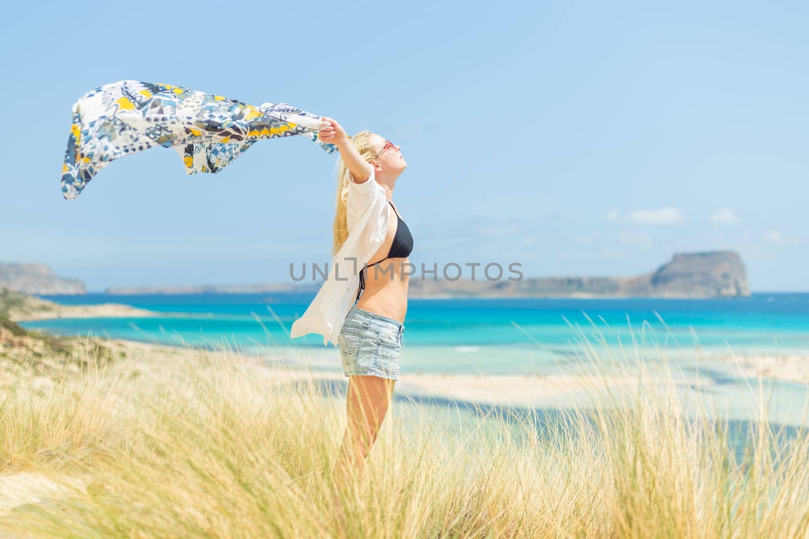 Relaxed woman, arms raised, holding colorful scarf, enjoying sun, freedom and life at beautiful beach. Young lady feeling free, relaxed and happy. Concept of vacations, freedom, joy and well being.