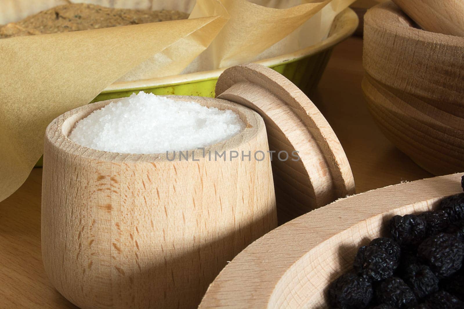 Close up of small wooden cup full of salt sitting on the kitchen table. Other kitchen wares blurred in the background