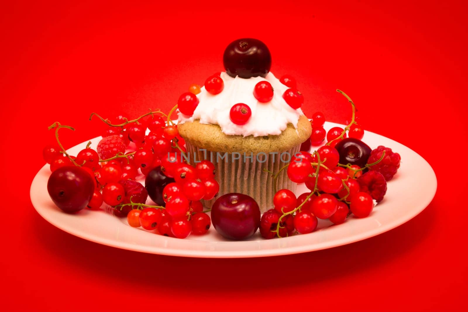 White plate with cupcake surrounded by red berries and cherries isolated on red background