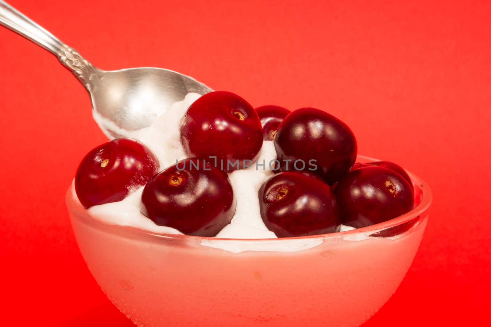 Picking up cherries with a spoon from a dessert in a glass cup on red background