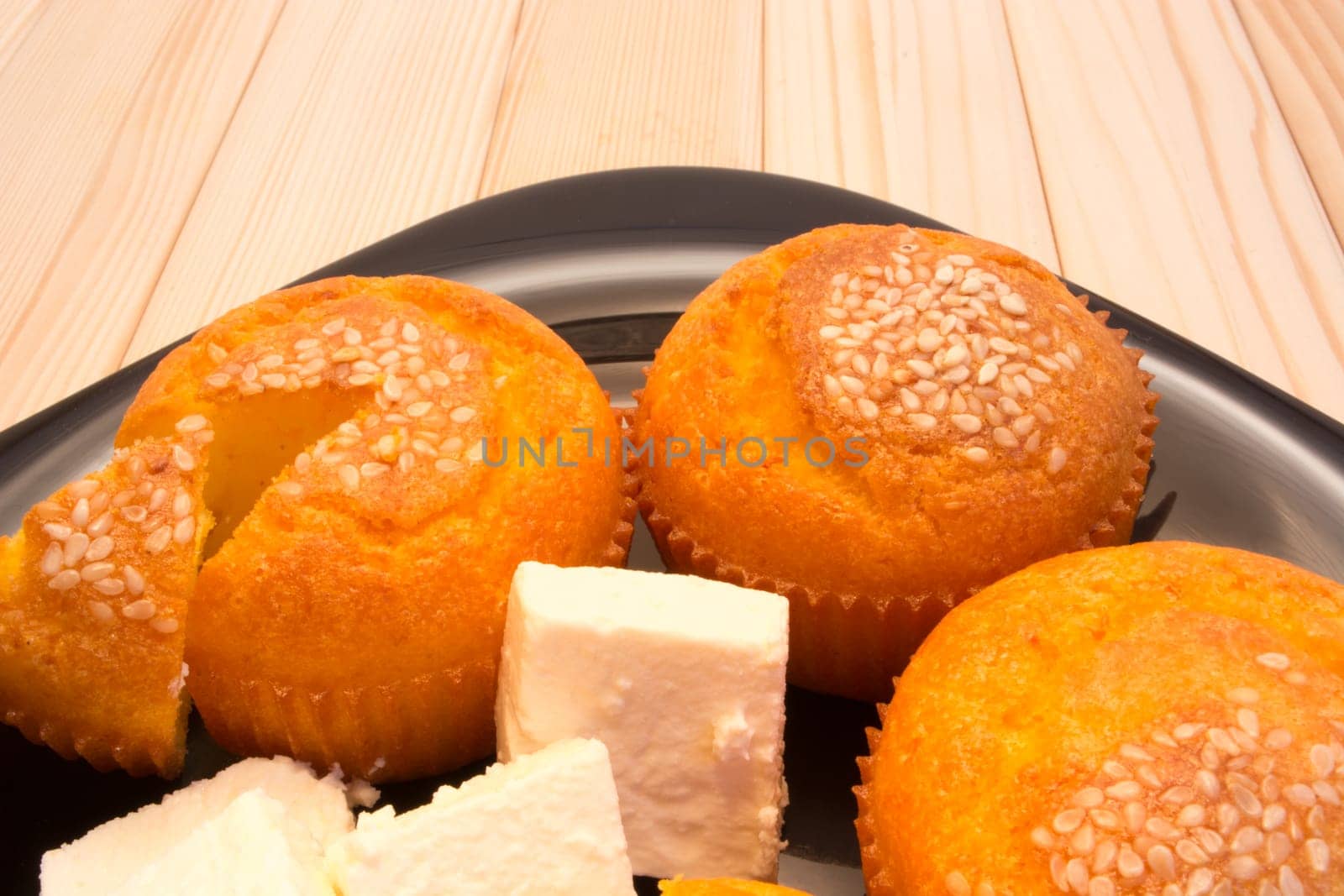 Black plate with muffins with sesame seeds on top on wooden table. Chops of cheese also on the plate